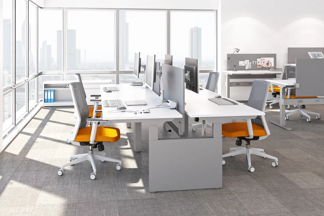 Studio photography of AMQ's KINEX Benching. A 2-Pack double run workspace is at the corner by the window. Another pair of single run is off to the side. Each has a white laminate table top, and adjustable screens.