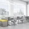 Global's 6-person power beam desks in an office space with tall ceilings. Each workstation has a laptop open, with glass paneling for privacy. Sunlight is cast upon the acoujou finish. At one end is a cabinet with sunflower yellow cushions, and a square Swap table.