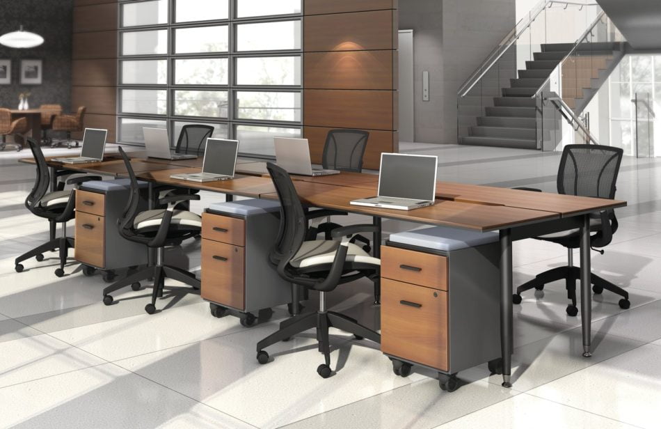 Studio photography of Global's 6-person sidebar benching with a vibrant honey brown finish. Each work station has mobile pedestal drawers and a rolling chair. Beyond, is a flight of stairs and elevator.