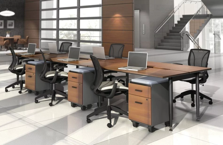 Studio photography of Global's 6-person sidebar benching with a vibrant honey brown finish. Each work station has mobile pedestal drawers and a rolling chair. Beyond, is a flight of stairs and elevator.