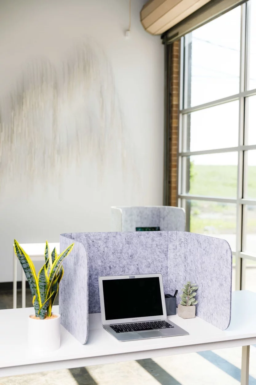 Hide privacy screen placed at a white desk. A smaller table is pushed up against the wall, by the window. It holds another workstation with Hide privacy screen.