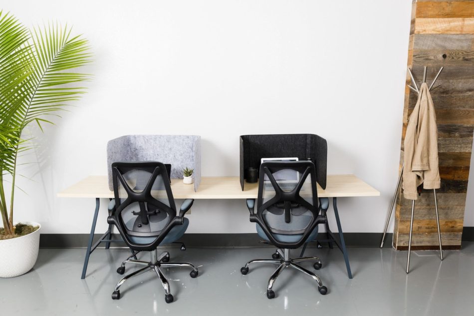 A wooden table against the wall of an open office space. Two office chairs are placed at the table, with a Hide model privacy screen. To the right, a tripod style coat rack holds a beige garment.