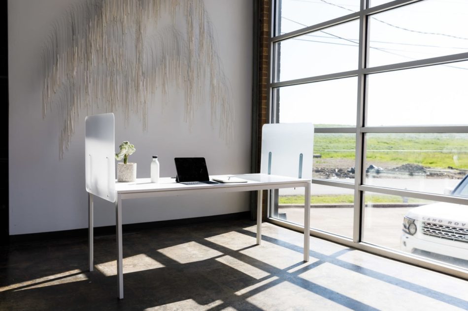 Simple table desk placed at the window in an office. Shelter screens lines the sides of the desk, and a laptop sits with its lid open.