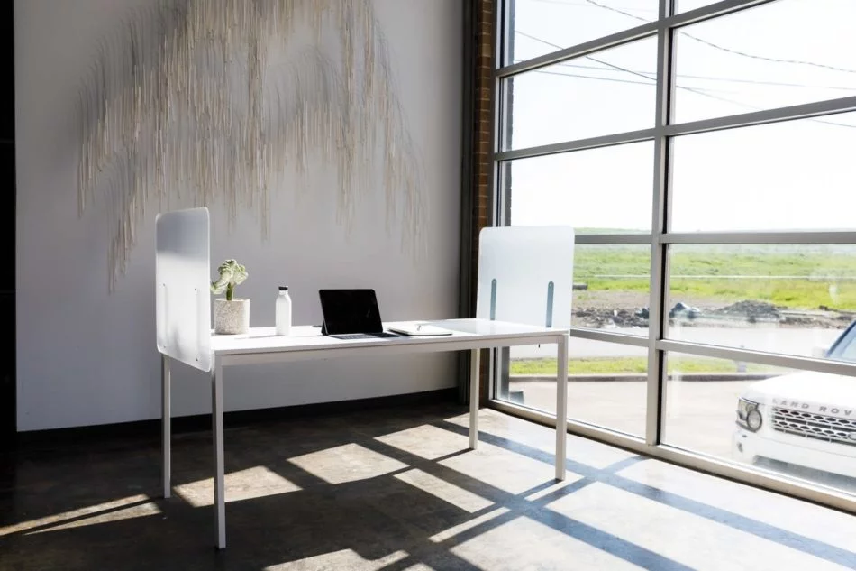 Simple table desk placed at the window in an office. Shelter screens lines the sides of the desk, and a laptop sits with its lid open.