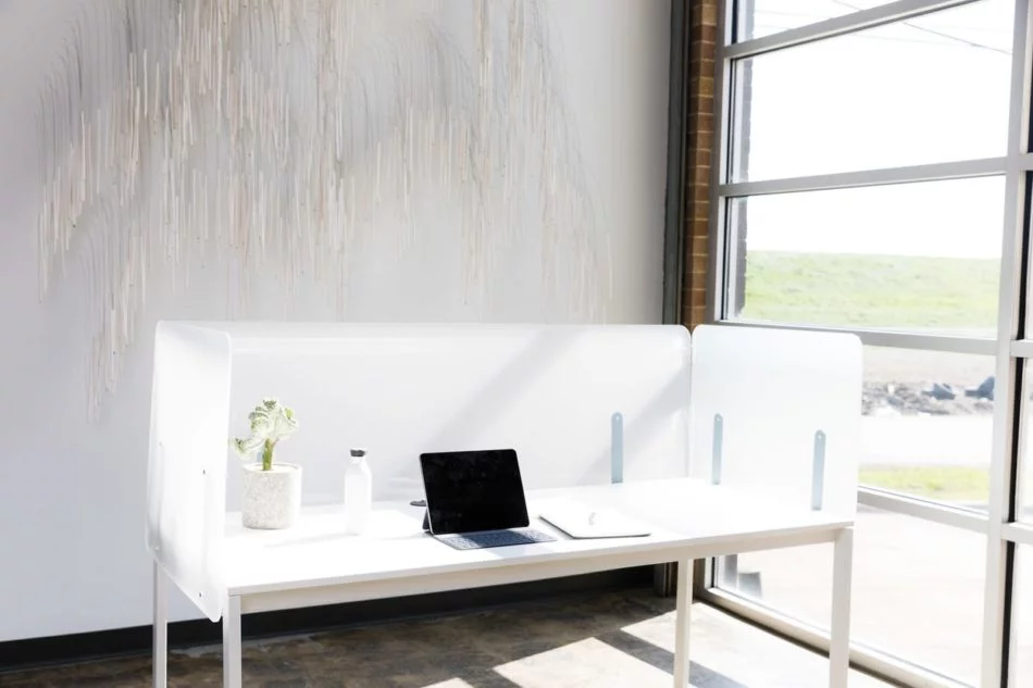 Simple table desk placed at the window in an office. Shelter screens line the desk, with an open laptop and notebook set on the table.