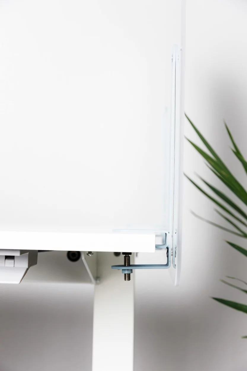 Closeup showing the edge of a desk, which has a Shelter acrylic screen added to it. You can see the screen's mounting bracket, showing from beneath the desk.