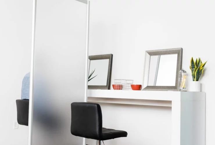 Two workstations side by side. A silver based stool is placed at each side, with a Split frosted acrylic screen in between. There is a framed mirror and plant resting at each work station.