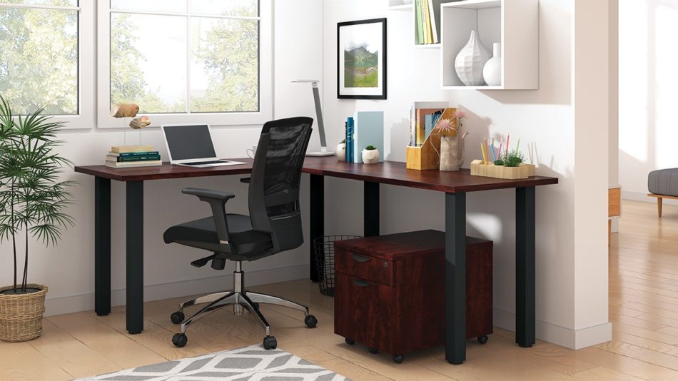 Studio shot of OTG's Home Office, with an L-shaped desk with a black colored metal frame. The surface is finished in an American Dark Cherry. A matchindg set of drawers has been rolled under the desk, at the left of the mesh back chair. To the left of the laptop is a set of books and an OTG LED Desk lamp in the corner.