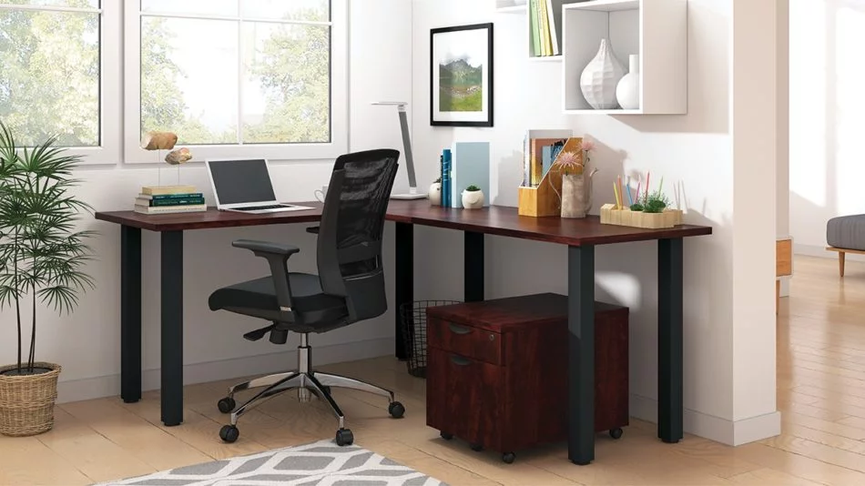 Studio shot of OTG's Home Office, with an L-shaped desk with a black colored metal frame. The surface is finished in an American Dark Cherry. A matchindg set of drawers has been rolled under the desk, at the left of the mesh back chair. To the left of the laptop is a set of books and an OTG LED Desk lamp in the corner.