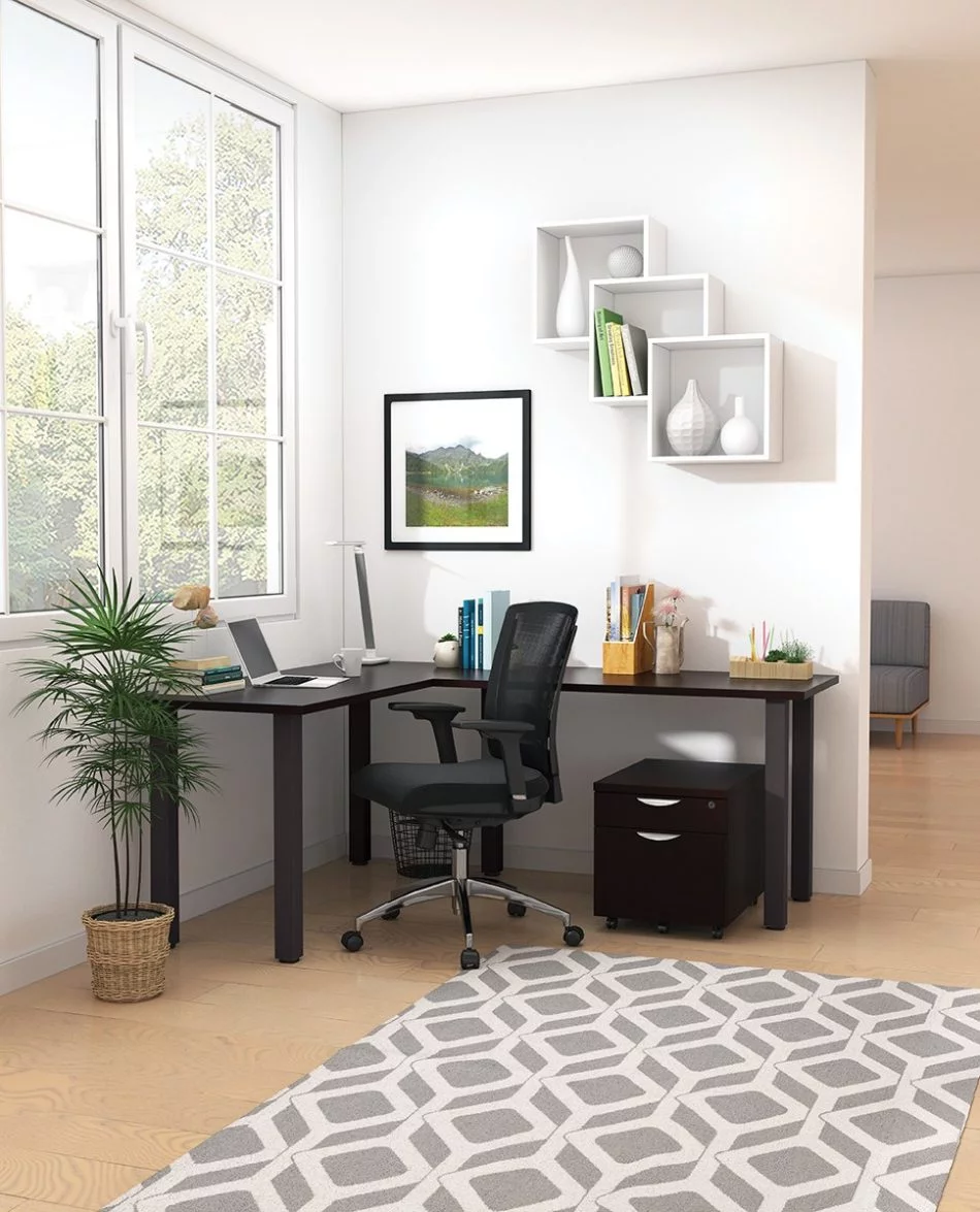 Studio shot of OTG's Home Office, with an L-shaped desk placed by the window. It has a black colored metal frame and top surfaces in an American Espresso finish. A matching set of drawers has been rolled under the desk, by a set of pencil cases. The office chair is seated by a laptop and an OTG LED Desk lamp in the corner.