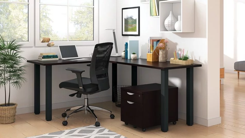 Studio shot of OTG's Home Office, with an L-shaped desk with a black colored metal frame. The surface is finished in American Espresso. A matching set of drawers has been rolled under the desk, at the left of the mesh back chair. To the left of the laptop is a set of books and an OTG LED Desk lamp in the corner.