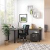 Studio shot of OTG's Home Office, with an L-shaped desk placed by the window. It has a black colored metal frame and top surfaces in an Artisan Grey finish. A matching set of drawers has been rolled under the desk, by a set of pencil cases. The office chair is seated by a laptop and an OTG LED Desk lamp in the corner.