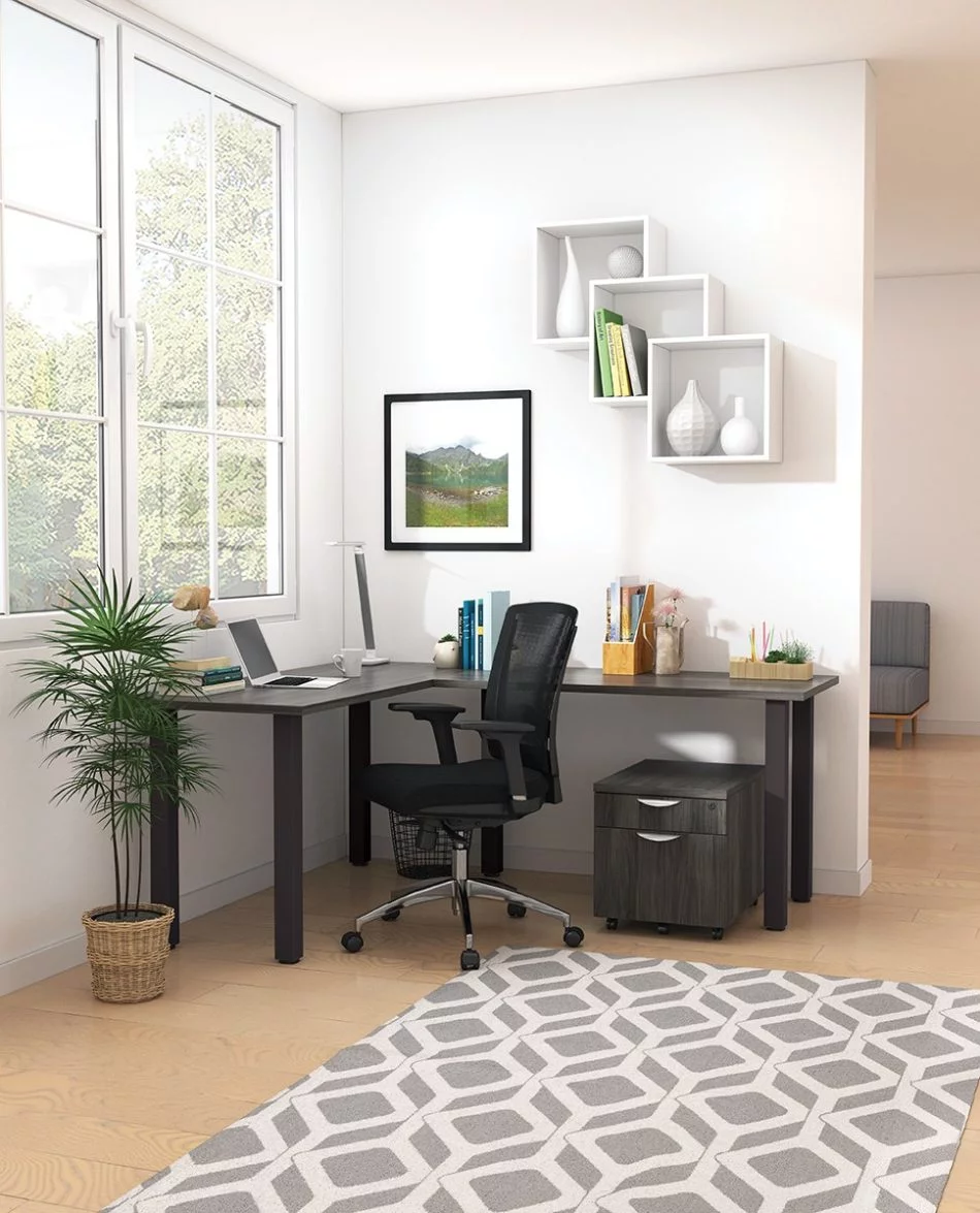 Studio shot of OTG's Home Office, with an L-shaped desk placed by the window. It has a black colored metal frame and top surfaces in an Artisan Grey finish. A matching set of drawers has been rolled under the desk, by a set of pencil cases. The office chair is seated by a laptop and an OTG LED Desk lamp in the corner.
