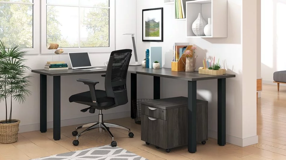 Studio shot of OTG's Home Office, with an L-shaped desk with a black, metal frame. The surface is finished in Artisan Grey. A matching set of drawers has been rolled under the desk, at the left of the mesh back chair. To the left of the laptop is a set of books and an OTG LED Desk lamp in the corner.