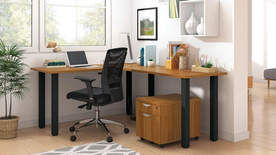 Studio shot of OTG's Home Office, with an L-shaped desk with a black colored metal frame. The surface is finished in Autumn Walnut. A matching set of drawers has been rolled under the desk, at the left of the mesh back chair. To the left of the laptop is a set of books and an OTG LED Desk lamp in the corner.