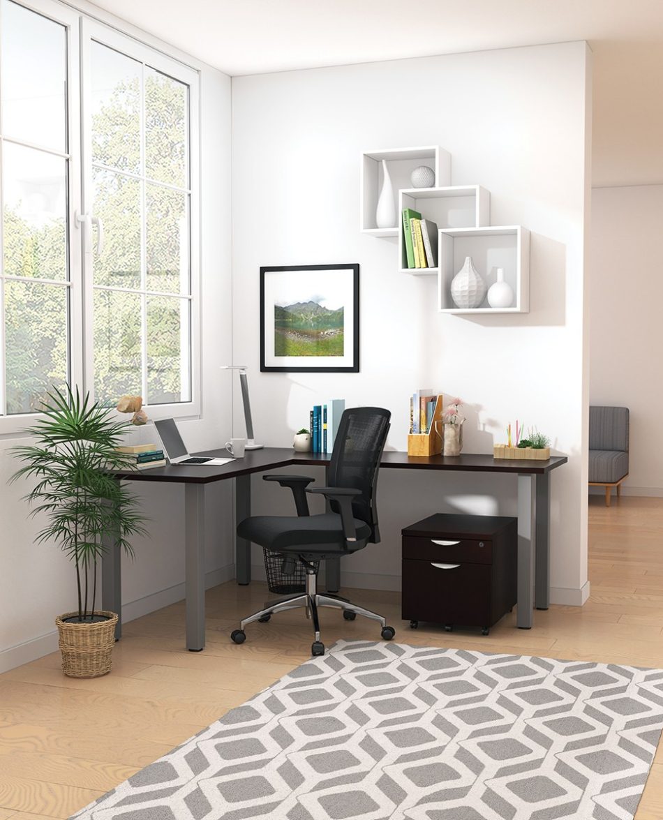 Studio shot of OTG's Home Office, with an L-shaped desk placed by the window. It has a tungsten colored metal frame and top surfaces in an American Espresso finish. A matching set of drawers has been rolled under the desk, by a set of pencil cases. The office chair is seated by a laptop and an OTG LED Desk lamp in the corner.