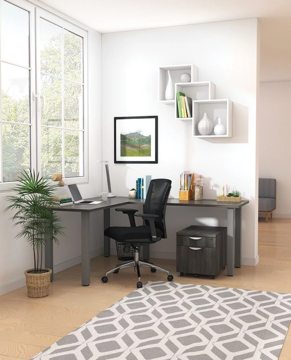 Studio shot of OTG's Home Office, with an L-shaped desk placed by the window. It has a tungsten colored metal frame and top surfaces in an Artisan Grey finish. A matching set of drawers has been rolled under the desk, by a set of pencil cases. The office chair is seated by a laptop and an OTG LED Desk lamp in the corner.