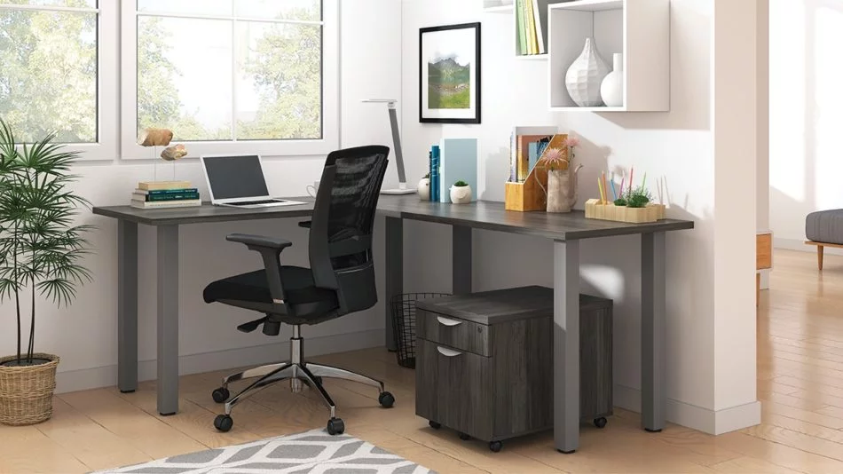 Studio shot of OTG's Home Office, with an L-shaped desk with a tungsten metal frame. The surface is finished in Artisan Grey. A matching set of drawers has been rolled under the desk, at the left of the mesh back chair. To the left of the laptop is a set of books and an OTG LED Desk lamp in the corner.