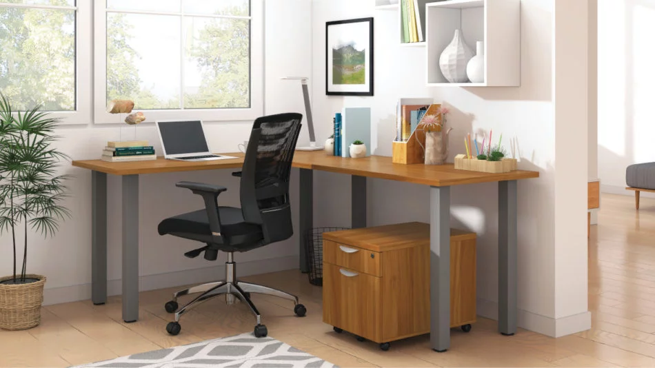 Studio shot of OTG's Home Office, with an L-shaped desk with a tungsten metal frame. The surface is finished in Autumn Walnut. A matching set of drawers has been rolled under the desk, at the left of the mesh back chair. To the left of the laptop is a set of books and an OTG LED Desk lamp in the corner.