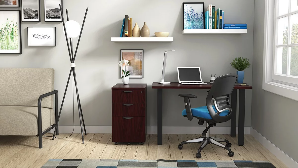 Studio shot of OTG's Home Office Setup, which has a 24" x 48" work table placed in the corner by the window. At the left is a set of pedestal drawers. The third (top) drawer has a lock on it. Both the desk and pedestal drawers are finished in an _____. A simple mesh-back office chair is pulled up to the laptop, with a LED lamp next to it.