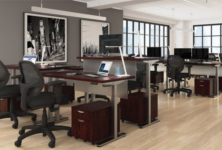 Studio photography of Offices to Go's laminate height adjustable workstations. Two groups of four stations are set together, each with an LED desk lamp and computer. A set of 2 drawers is wheeled in each space, with storage for supplies. Sunlight comes in from windows in the back of this photo. A pair of large black and white paintings hang on a nearby wall.