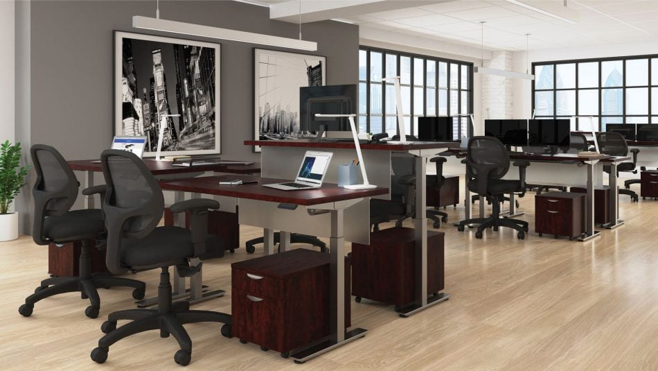 Studio photography of Offices to Go's laminate height adjustable workstations. Two groups of four stations are set together, each with an LED desk lamp and computer. A set of 2 drawers is wheeled in each space, with storage for supplies. Sunlight comes in from windows in the back of this photo. A pair of large black and white paintings hang on a nearby wall.