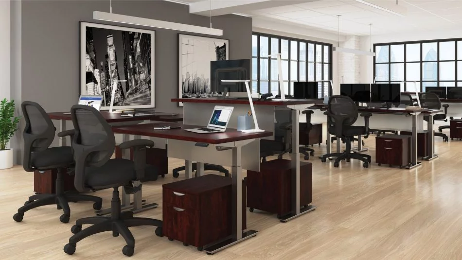 Studio photography of Offices to Go's laminate height adjustable workstations. Two groups of four stations are set together, each with an LED desk lamp and computer. A set of 2 drawers is wheeled in each space, with storage for supplies. Sunlight comes in from windows in the back of this photo. A pair of large black and white paintings hang on a nearby wall.