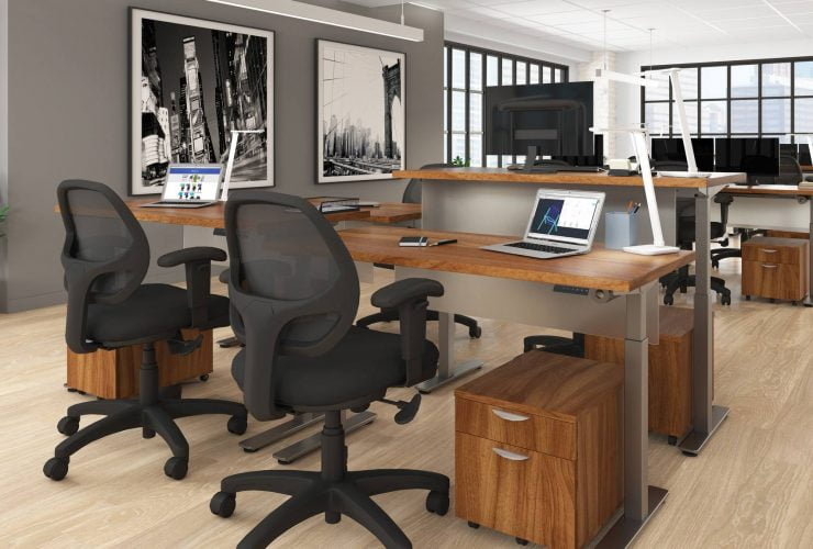 Studio photography of Offices to Go's laminate height adjustable workstations. A group of three stations are set together, each with an LED desk lamp and computer. A set of 2 drawers is wheeled in each space, with storage for supplies. Sunlight comes in from windows towards the background. A pair of large black and white paintings hang on the wall off to the right.