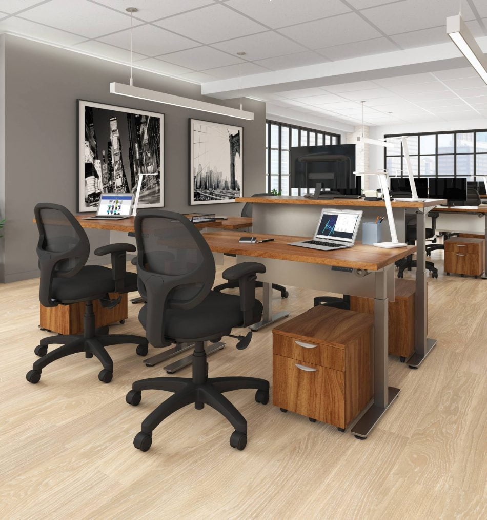 Studio photography of Offices to Go's laminate height adjustable workstations. A group of three stations are set together, each with an LED desk lamp and computer. A set of 2 drawers is wheeled in each space, with storage for supplies. Sunlight comes in from windows towards the background. A pair of large black and white paintings hang on the wall off to the right.