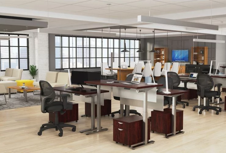 Studio photography of Offices to Go's laminate height adjustable workstations. Two groups of four stations are set together, each with an LED desk lamp and computer. A set of 2 drawers is wheeled in each space, with storage for supplies. A clear glass conference room can be seen in the background, by the open office's windows, with an additional casual seating area to the left.