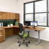 Studio photography of an Offices to Go furniture set, placed in the corner. The credenza and height adjustable table are placed in an L-formation. An overhead hutch attaches to the credenza, with some picture frames placed on the table top. Dual monitors are placed along the height adjustable extension, with a high backed chair facing the window,