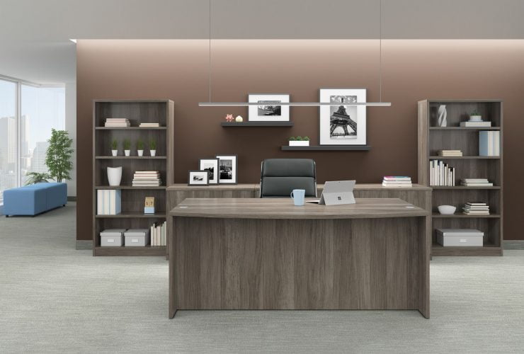 Studio photography of Offices to Go furniture in an open executive or management space. A bow front desk holds a laptop and mobile accessory, with a black mid-back pleather chair placed behind it. The room is lit from a recessed area above, adding more light to the wall behind. An extra wide mixed storage unit is between two tall bookcases. Some shelves are placed above the storage unit, with photography and art. The bookcase, mixed storage unit and desk use an Artisan Grey laminate finish.