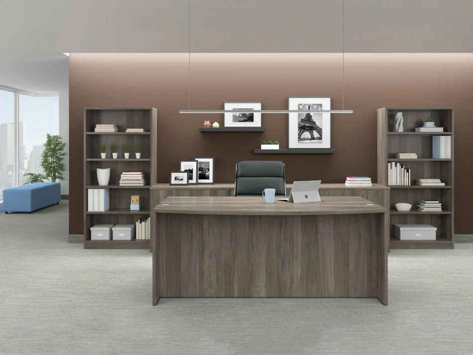 Studio photography of Offices to Go furniture in an open executive or management space. A bow front desk holds a laptop and mobile accessory, with a black mid-back pleather chair placed behind it. The room is lit from a recessed area above, adding more light to the wall behind. An extra wide mixed storage unit is between two tall bookcases. Some shelves are placed above the storage unit, with photography and art. The bookcase, mixed storage unit and desk use an Artisan Grey laminate finish.