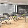 Studio photography of Offices to Go's laminate flip-top training tables. Six of these tables have been placed in front of a single 4-legged laminate table for the instructor. Each classroom table has a tungsten colored frame, rolling casters, and an American Espresso finish on the tabletop. At each training table are two tungsten framed nesting chairs, with a plush lime green seat cushion. Windows at the back of the room, show a downtown area beyond.