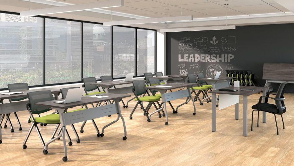 Studio photography of Offices to Go's laminate flip-top training tables. Six of these tables have been placed in front of a single 4-legged laminate table for the instructor. Each classroom table has a tungsten colored frame, rolling casters, and an American Espresso finish on the tabletop. At each training table are two tungsten framed nesting chairs, with a plush lime green seat cushion. Windows at the back of the room, show a downtown area beyond.