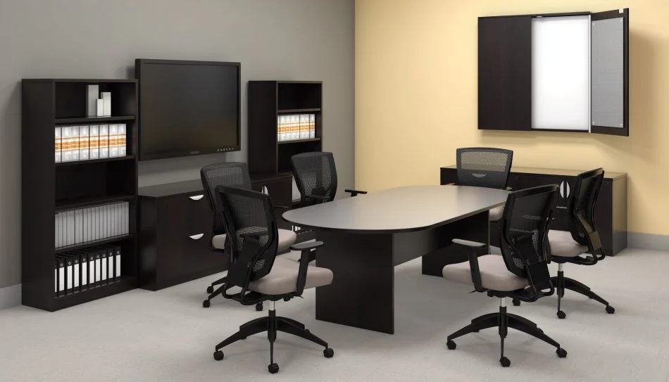 Studio photography of Offices to Go furniture in a conference area. A racetrack shaped table is in the center, with 6 high-back chairs placed around it. On the far wall, a presentation board has one of its double doors open, revealing a white board underneath. At the left is a matching mixed storage unit, with a tall book case at each side, and a flat screen TV directly above. The furniture uses an American Espresso finish.