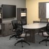 Studio photography of Offices to Go furniture in a conference area. A racetrack shaped table is in the center, with 6 high-back chairs placed around it. On the far wall, a presentation board has one of its double doors open, revealing a white board underneath. At the left is a matching mixed storage unit, with a tall book case at each side, and a flat screen TV directly above. The furniture uses an Artisan Grey finish.