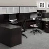 Studio photography of Offices to Go furniture, consisting of two L shape desks against an outside corner wall. There are two chairs and a laptop at each workstation. Overhead is a storage hutch that has silver double doors. This desk uses an American Espresso (laminate) finish. In the background is another desk for reception, by the window.