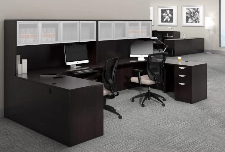 Studio photography of Offices to Go furniture, consisting of two L shape desks against an outside corner wall. There are two chairs and a laptop at each workstation. Overhead is a storage hutch that has silver double doors. This desk uses an American Espresso (laminate) finish. In the background is another desk for reception, by the window.