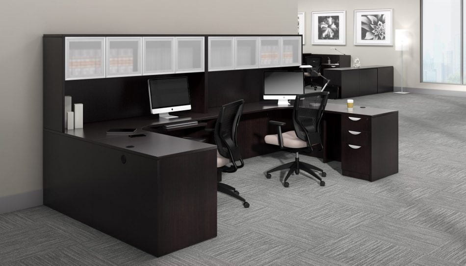 Studio photography of Offices to Go furniture, consisting of two L shape desks against an outside corner wall. There are two chairs and a laptop at each workstation. Overhead is a storage hutch that has silver double doors. This desk uses an American Espresso (laminate) finish. In the background is another desk for reception, by the window.