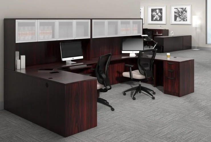 Studio photography of Offices to Go furniture, consisting of two L shape desks against an outside corner wall. There are two chairs and a laptop at each workstation. Overhead is a storage hutch that has silver double doors. This desk uses an American Mahogany (laminate) finish. In the background is another desk for reception, by the window.
