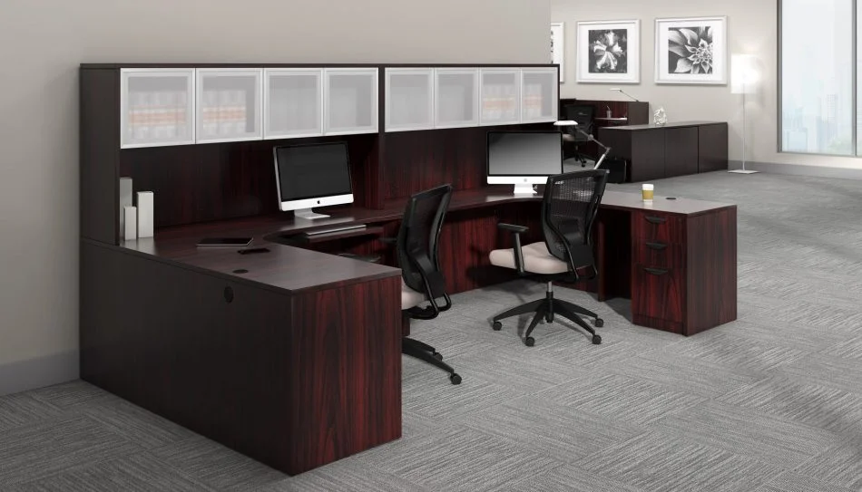 Studio photography of Offices to Go furniture, consisting of two L shape desks against an outside corner wall. There are two chairs and a laptop at each workstation. Overhead is a storage hutch that has silver double doors. This desk uses an American Mahogany (laminate) finish. In the background is another desk for reception, by the window.
