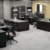 Studio photography of an Offices to Go furniture, set up in an open office near the elevator. Two L shaped desks are pressed against one another, giving a wide surface area for those passing in front. Another set like this is reversed, facing the far back wall. A large credenza exists in the wide area between these work stations. This office's desking uses an American Espresso finish.