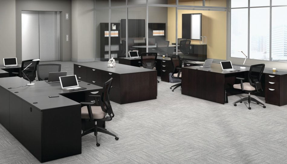 Studio photography of an Offices to Go furniture, set up in an open office near the elevator. Two L shaped desks are pressed against one another, giving a wide surface area for those passing in front. Another set like this is reversed, facing the far back wall. A large credenza exists in the wide area between these work stations. This office's desking uses an American Espresso finish.