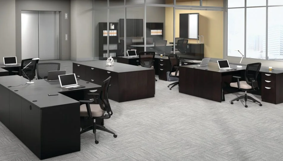Studio photography of an Offices to Go furniture, set up in an open office near the elevator. Two L shaped desks are pressed against one another, giving a wide surface area for those passing in front. Another set like this is reversed, facing the far back wall. A large credenza exists in the wide area between these work stations. This office's desking uses an American Espresso finish.