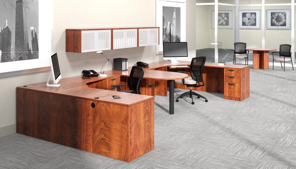 Studio photography of Offices to Go furniture, consisting of two L shape desks arranged from front to back, with a D-island just in between. There are two chairs pulled up to these L-shape workstations, a set of three overhead compartments mounted on the wall above. The furniture and shelving uses an American Dark Cherry finish.