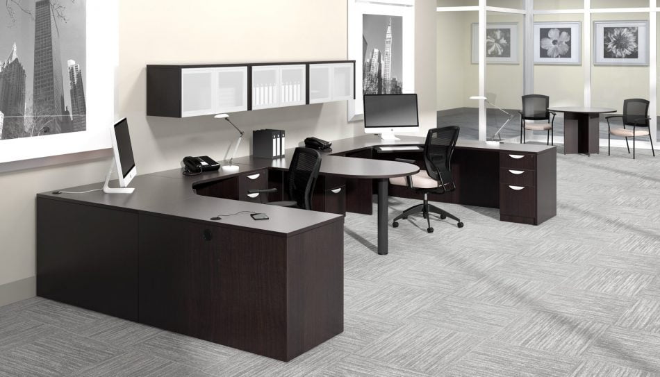 Studio photography of Offices to Go furniture, consisting of two L shape desks arranged from front to back, with a D-island just in between. There are two chairs pulled up to these L-shape workstations, a set of three overhead compartments mounted on the wall above. The furniture and shelving uses an American Espresso finish.