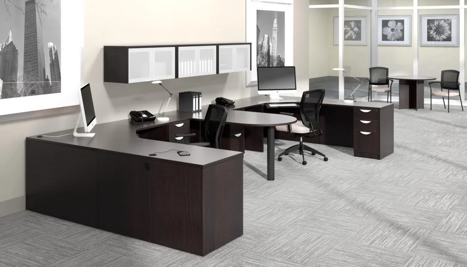 Studio photography of Offices to Go furniture, consisting of two L shape desks arranged from front to back, with a D-island just in between. There are two chairs pulled up to these L-shape workstations, a set of three overhead compartments mounted on the wall above. The furniture and shelving uses an American Espresso finish.