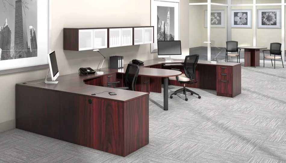 Studio photography of Offices to Go furniture, consisting of two L shape desks arranged from front to back, with a D-island just in between. There are two chairs pulled up to these L-shape workstations, a set of three overhead compartments mounted on the wall above. The furniture and shelving uses an American Mahogany finish.