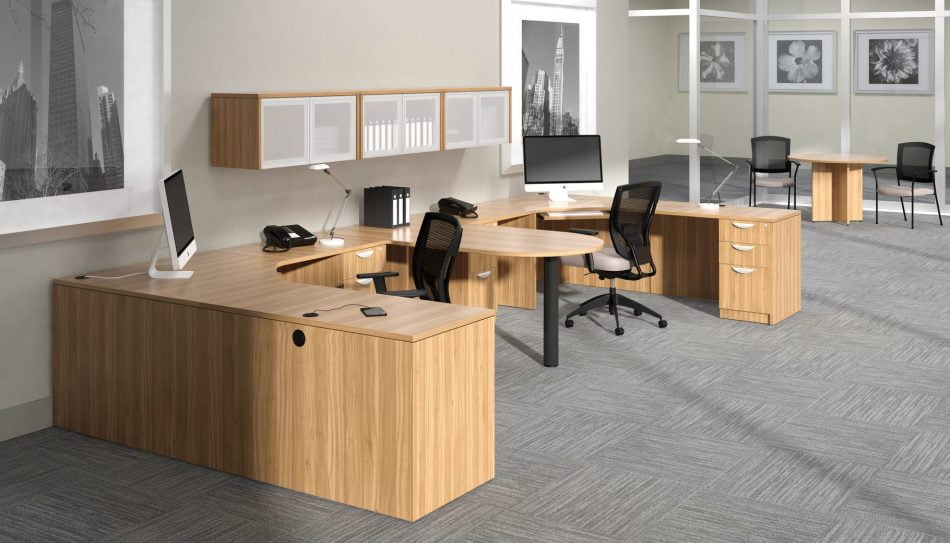 Studio photography of Offices to Go furniture, consisting of two L shape desks arranged from front to back, with a D-island just in between. There are two chairs pulled up to these L-shape workstations, a set of three overhead compartments mounted on the wall above. The furniture and shelving uses an Autumn Walnut finish.